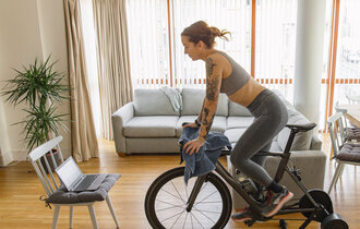 Eine Frau trainiert Zuhause auf einem Fahrrad Ergometer und hat dabei den Laptop aufgeklappt.