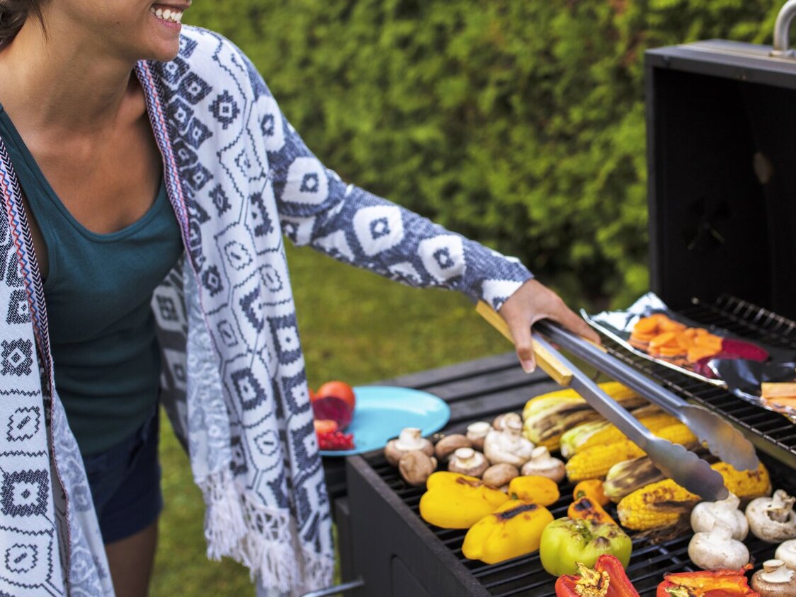 Eine Frau, die in einem Garten Gemüse grillt. Sie wendet es mit einer Zange und lächelt freudig dabei.