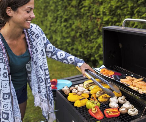 Eine Frau, die in einem Garten Gemüse grillt. Sie wendet es mit einer Zange und lächelt freudig dabei.