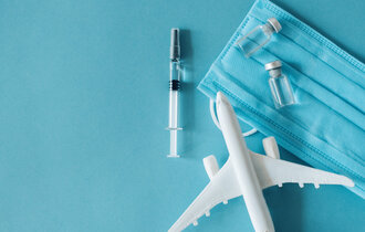 Auf blauem Hintergrund liegt ein Spielzeug-Flugzeug, eine Spritze mit Ampullen und ein Mundschutz.