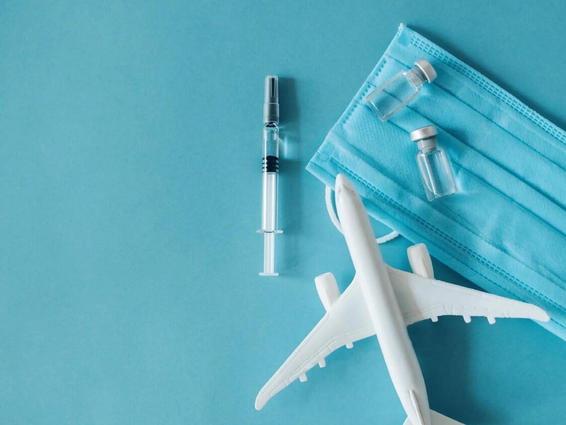 Auf blauem Hintergrund liegt ein Spielzeug-Flugzeug, eine Spritze mit Ampullen und ein Mundschutz.