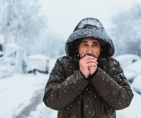 Mann läuft im Winter im Schnee eine Straße entlang, hat seine Kapuze auf und bläst sich in die Hände.