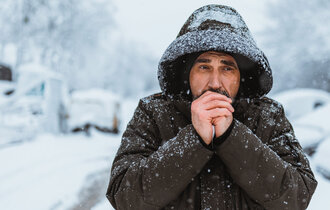 Mann läuft im Winter im Schnee eine Straße entlang, hat seine Kapuze auf und bläst sich in die Hände.