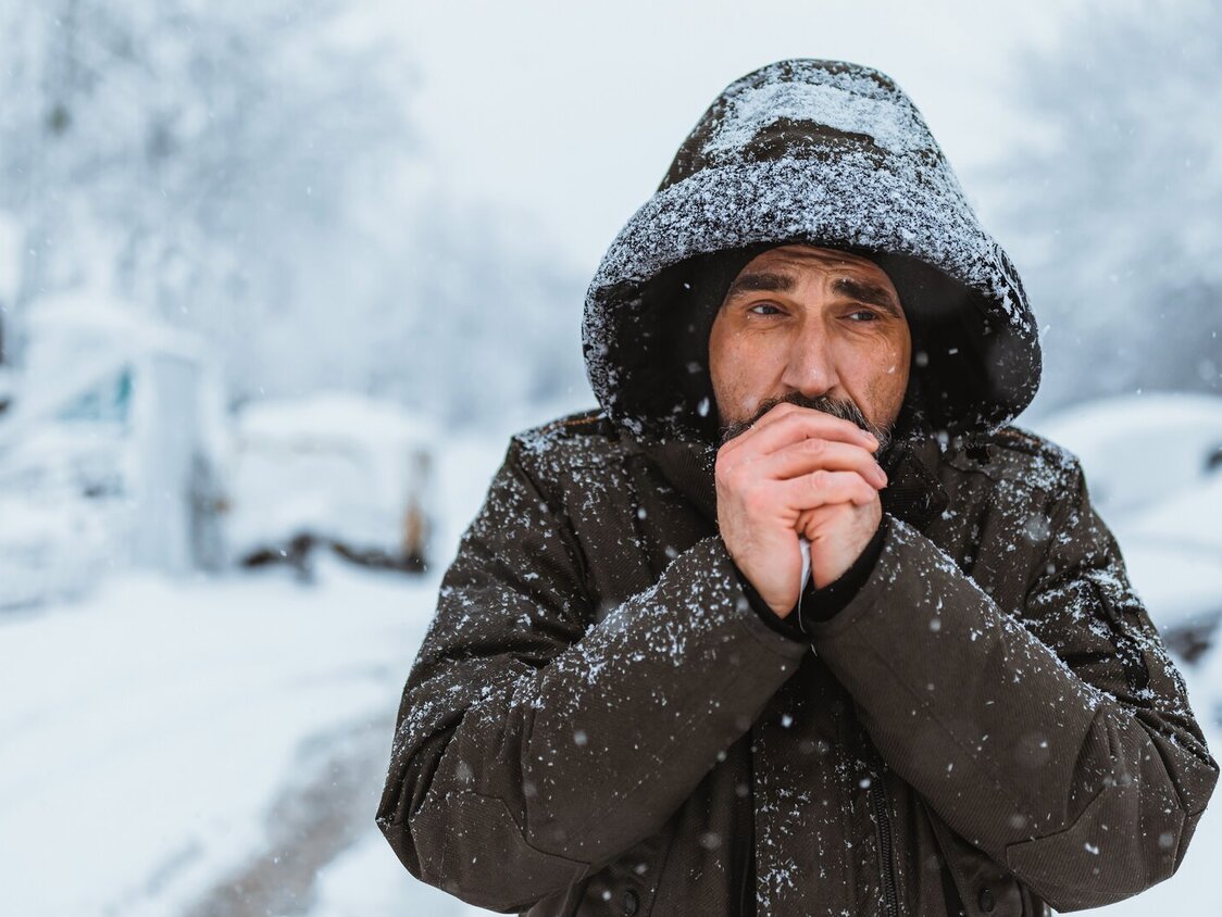 Mann läuft im Winter im Schnee eine Straße entlang, hat seine Kapuze auf und bläst sich in die Hände.