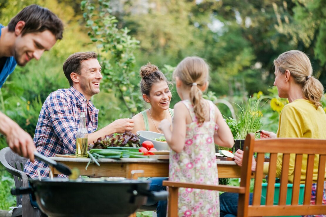 Eine Familie sitzt im Garten am Tisch. Eine Person steht davor am Grill.