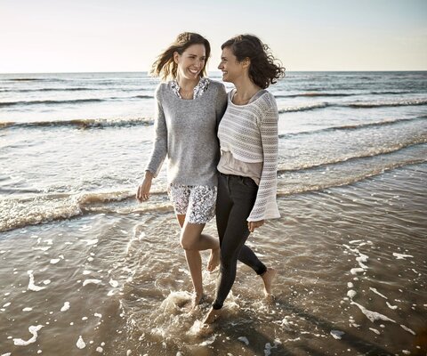 Zwei Freundinnen gehen Arm in Arm am Strand spazieren.
