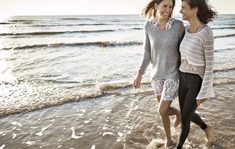 Zwei Freundinnen gehen Arm in Arm am Strand spazieren.