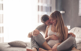 Ein Mann sitzt hinter seiner schwangeren Frau auf dem Bett und küsst ihren Hals.