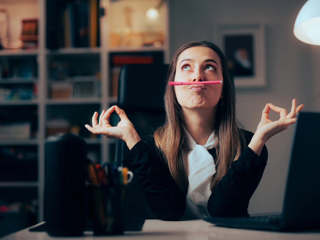 Frau im Büro balanciert Stift mit dem Mund und prokrastiniert.