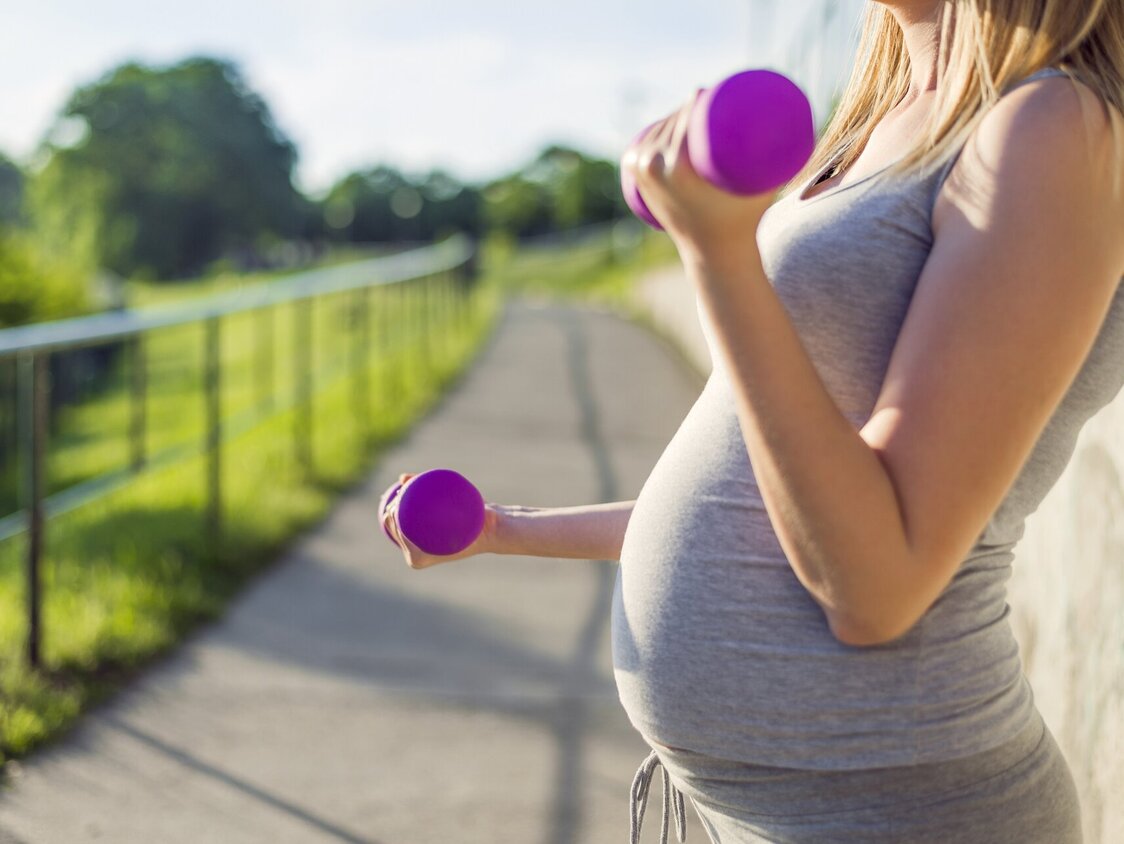 Eine schwangere Frau steht in Sportbekleidung draußen und hat kleine Hanteln in den Händen