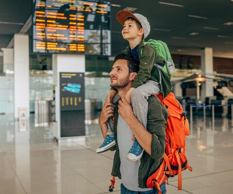 Ein Mann trägt einen Jungen Huckepack am Flughafen und hat dabei noch einen Rucksack auf.