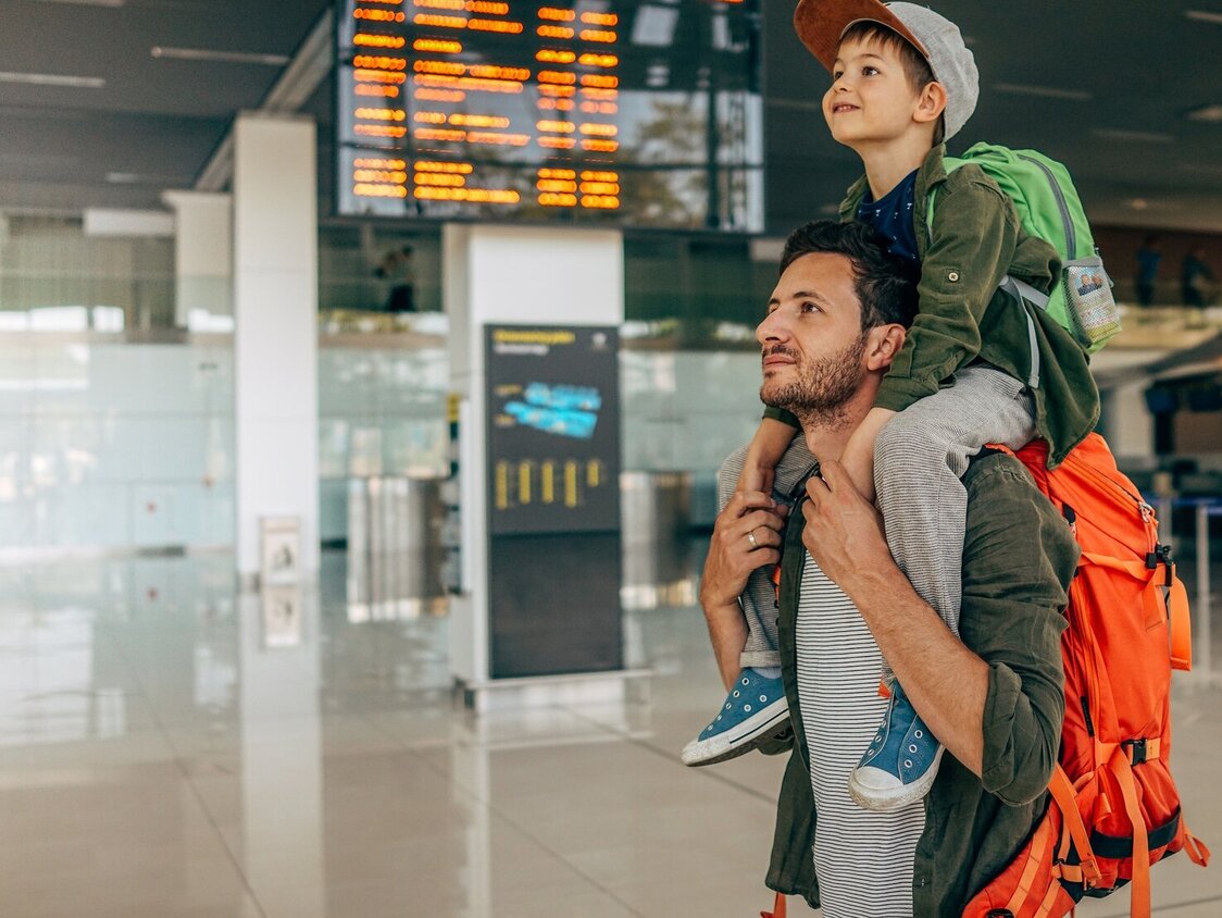 Ein Mann trägt einen Jungen Huckepack am Flughafen und hat dabei noch einen Rucksack auf.