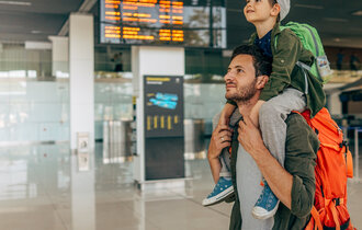 Ein Mann trägt einen Jungen Huckepack am Flughafen und hat dabei noch einen Rucksack auf.