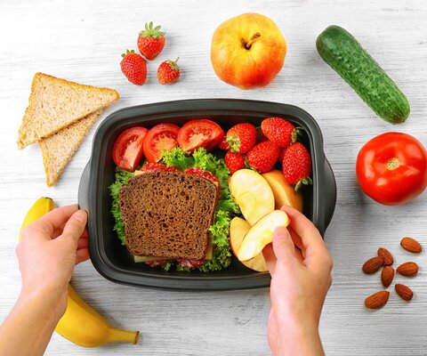 Eine Box mit einem frischen Brot mit Salat, Apfelschnitzen. Tomaten und Erdbeeren . Darum herum auch frisches Obst und Gemüse. Eine Hand greift einen Schnitz heraus.