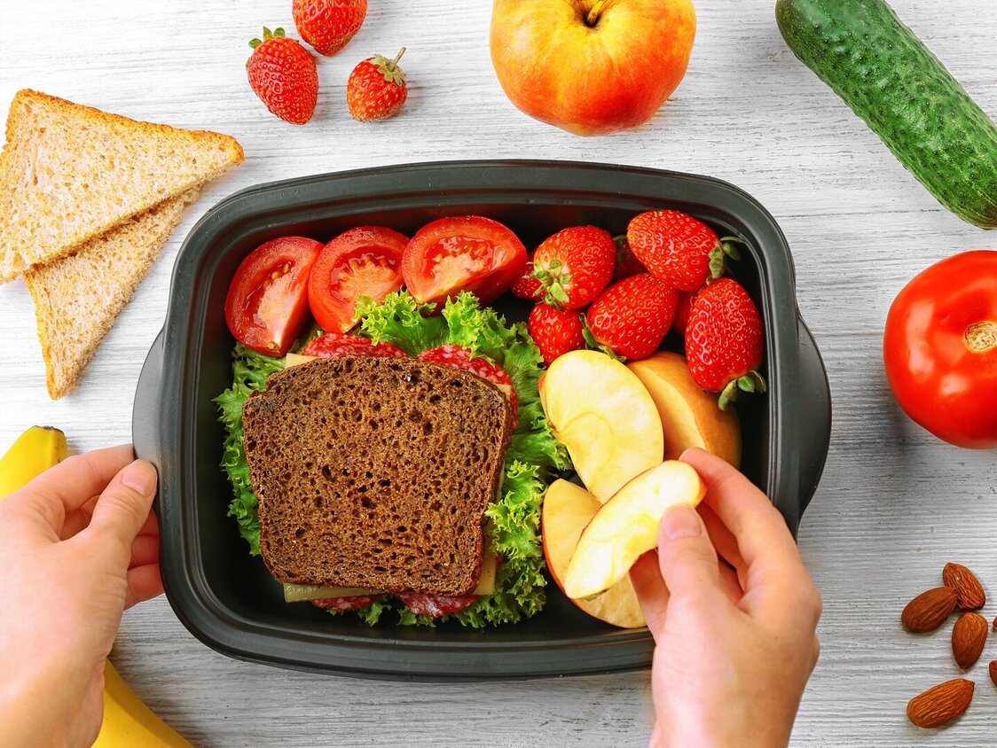 Eine Box mit einem frischen Brot mit Salat, Apfelschnitzen. Tomaten und Erdbeeren . Darum herum auch frisches Obst und Gemüse. Eine Hand greift einen Schnitz heraus.