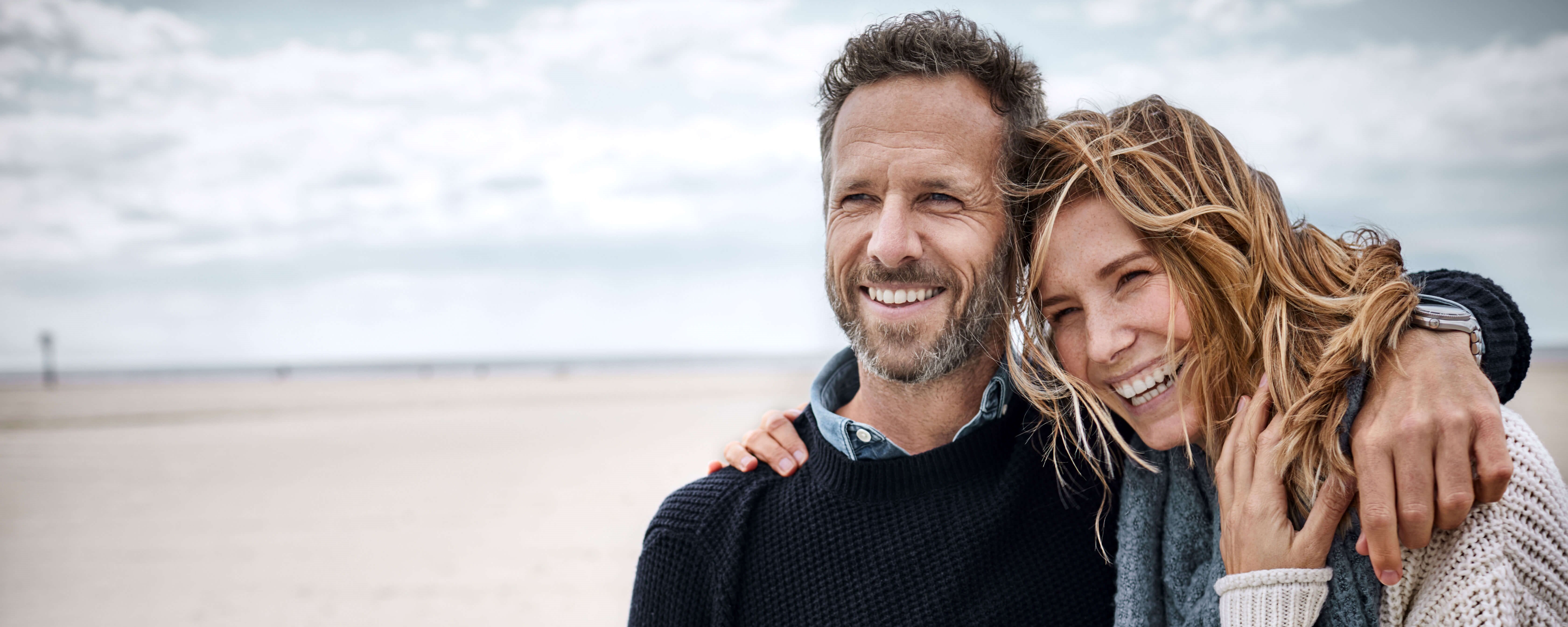 Ein Mann und eine Frau, Arm in Arm an Strand. Im Hintergrund eine Sanddüne, blauer Himmel und einige Wolken.