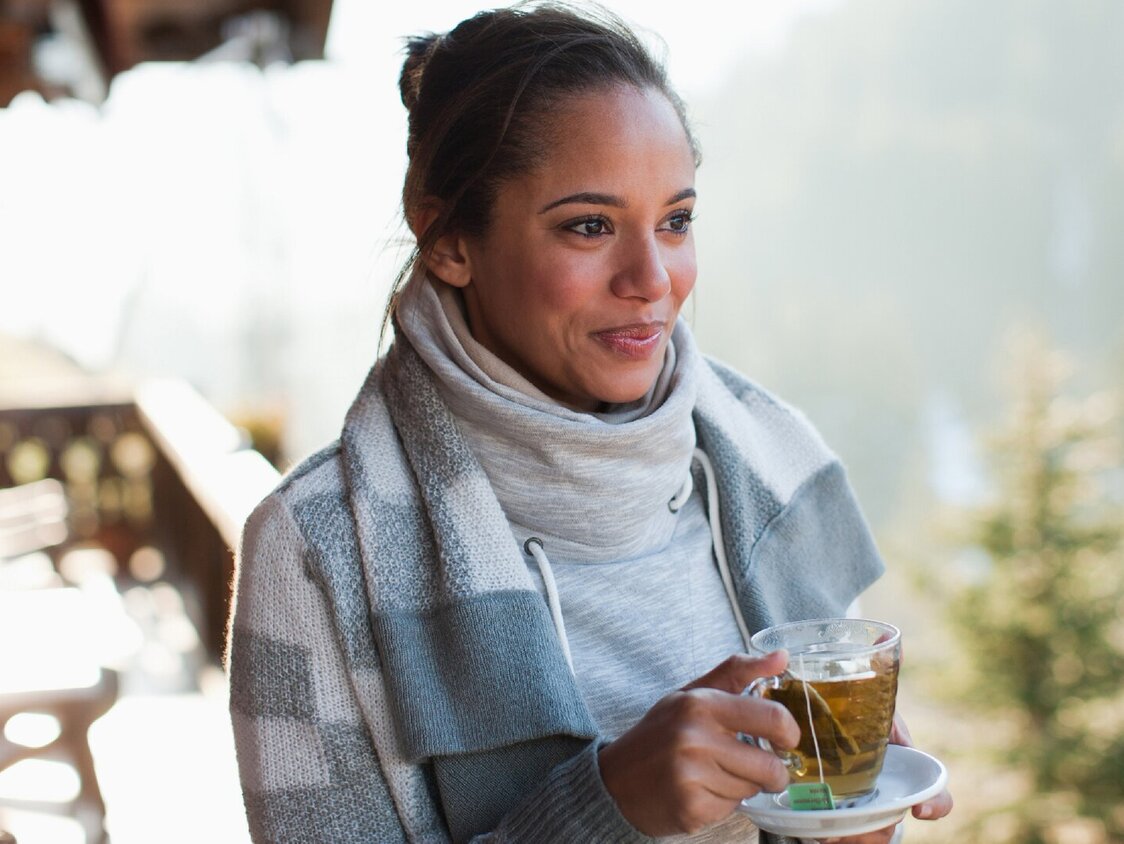 Eine lächelnde Frau trinkt draußen im Stehen einen Tee.