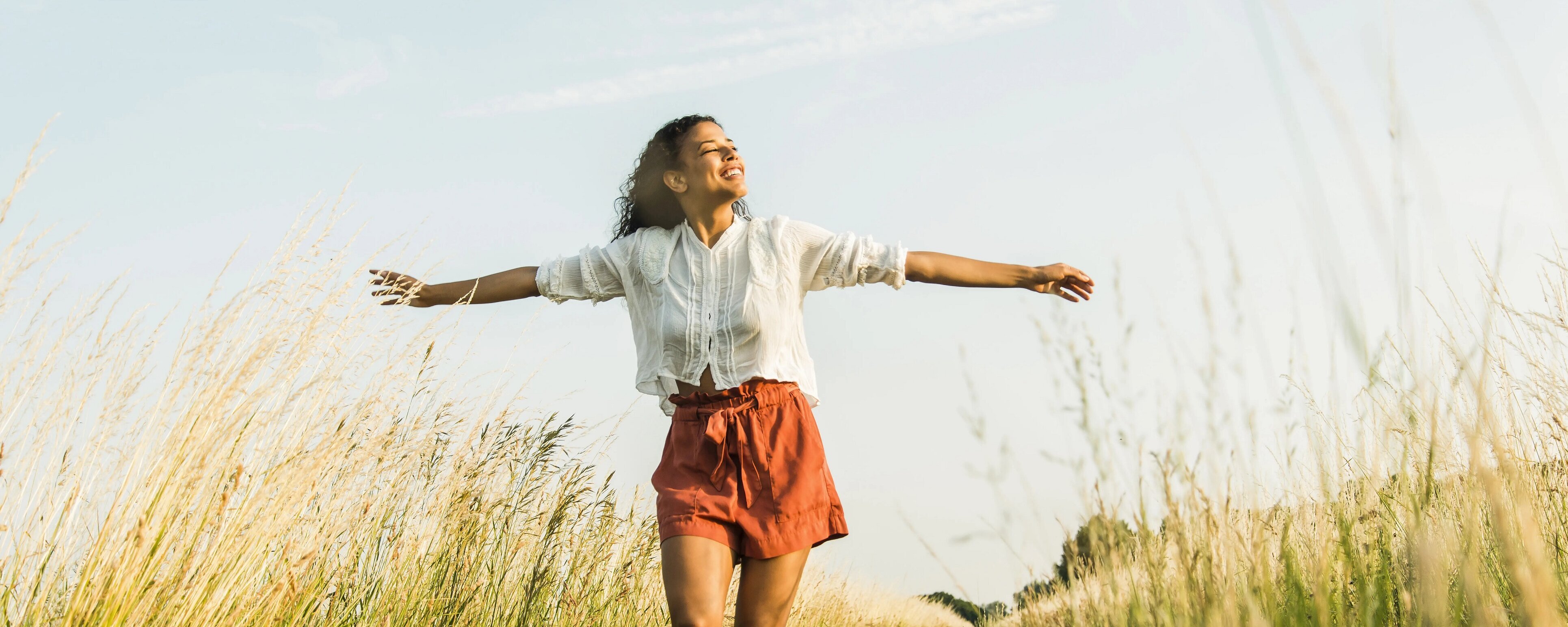 Eine Frau welche durch ein Feld oder über eine Wiese spaziert. Sie hält die Arme ausgebreitet und wendet ihr Gesicht der Sonne zu.