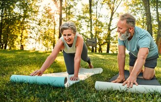 Ein älterer Mann und eine ältere Frau breiten auf einer Wiese ihre Yoga-Matten aus.