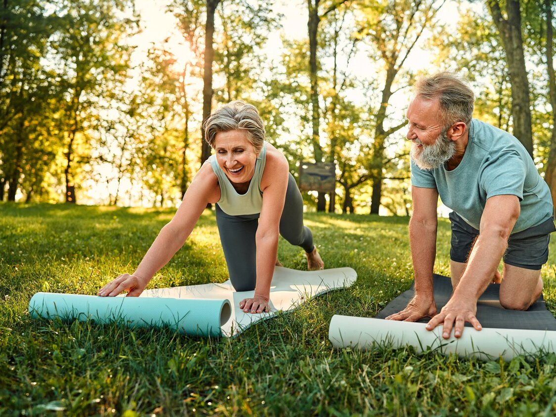 Ein älterer Mann und eine ältere Frau breiten auf einer Wiese ihre Yoga-Matten aus.