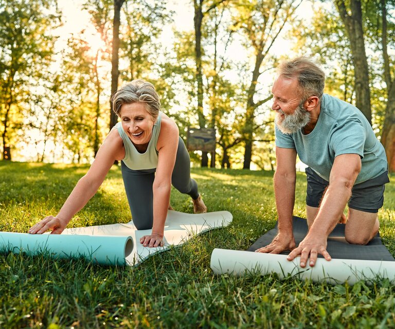 Ein älterer Mann und eine ältere Frau breiten auf einer Wiese ihre Yoga-Matten aus.