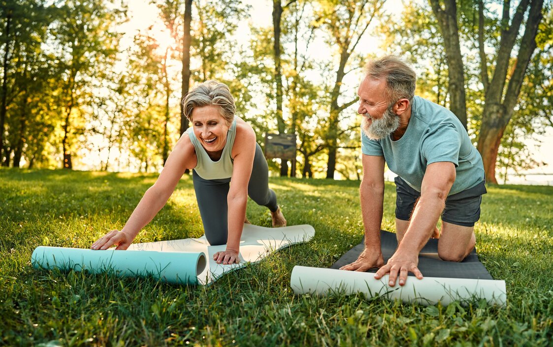 Ein älterer Mann und eine ältere Frau breiten auf einer Wiese ihre Yoga-Matten aus.