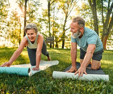 Ein älterer Mann und eine ältere Frau breiten auf einer Wiese ihre Yoga-Matten aus.