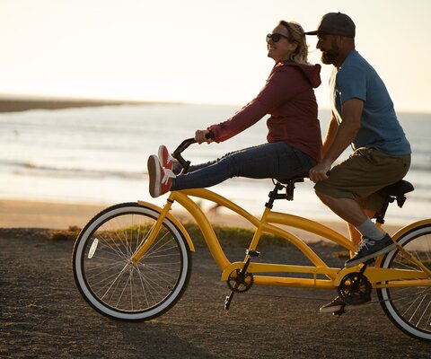An einem Strand fährt ein Paar mit einem gelben Tandem an der Promenade entlang.