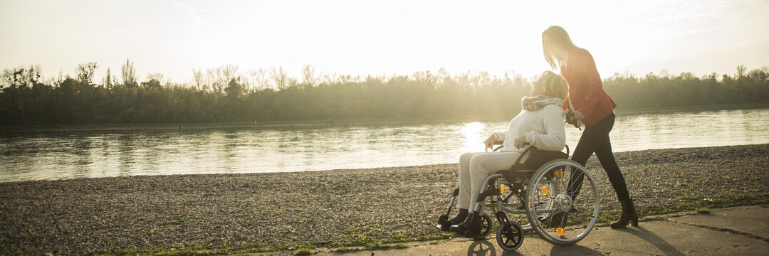 Eine Frau schiebt eine ältere Damen im Rollstuhl an einem Fluss entlang.
