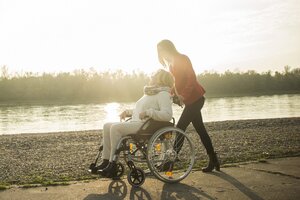 Eine Frau schiebt eine ältere Damen im Rollstuhl an einem Fluss entlang.