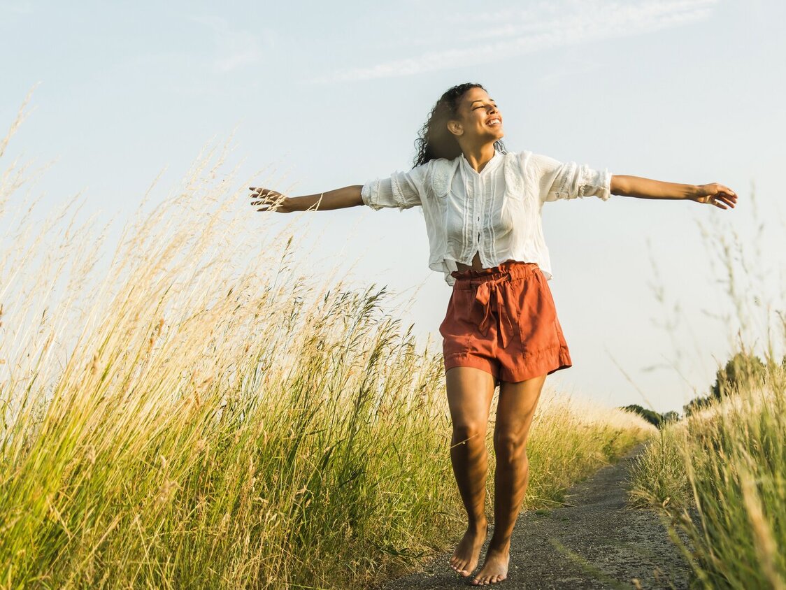 Eine junge Frau steht barfuß auf einem Weg in einem Feld und steckt ihre Arme aus.