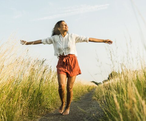 Eine junge Frau steht barfuß auf einem Weg in einem Feld und steckt ihre Arme aus.