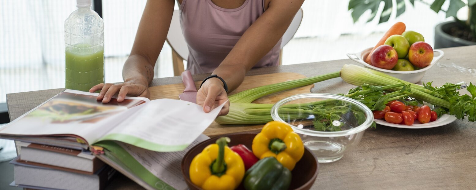 Eine asiatische Frau steht an einem Tisch mit einem aufgeschlagenen Kochbuch und drum herum liegt Gemüse.