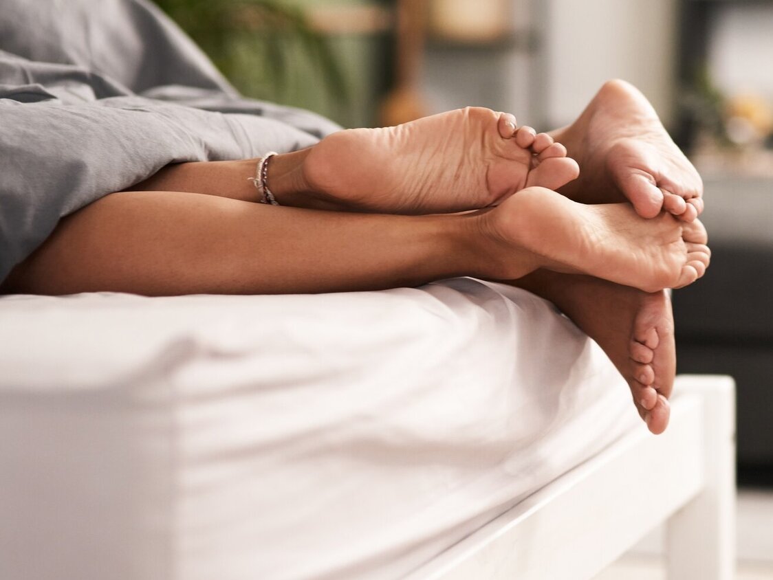 Ein Paar liegt im Bett und es gucken nur die Füße raus, die ineinander verschränkt sind.
