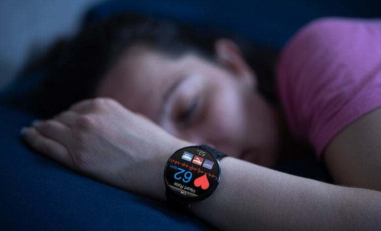 Eine digitale Uhr am Handgelenk einer schlafenden Frau. Die Uhr misst den Puls, symbolisiert durch ein Herz.