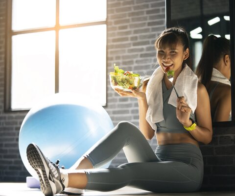 Eine Frau sitzt in einem Sportraum und isst einen Salat.