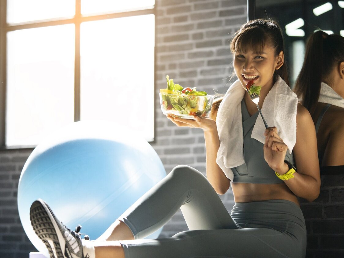 Eine Frau sitzt in einem Sportraum und isst einen Salat.