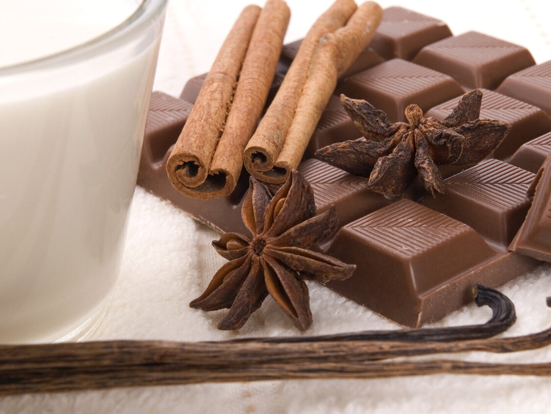 Neben einem Glas Milch liegt eine Tafel Schokolade, zwei Vanillestangen, zwei Stück Sternanis und ein paar Zimtstangen.