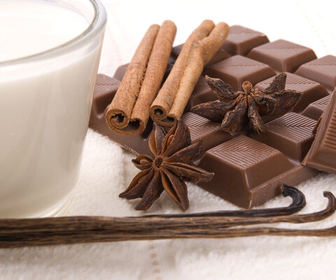 Neben einem Glas Milch liegt eine Tafel Schokolade, zwei Vanillestangen, zwei Stück Sternanis und ein paar Zimtstangen.
