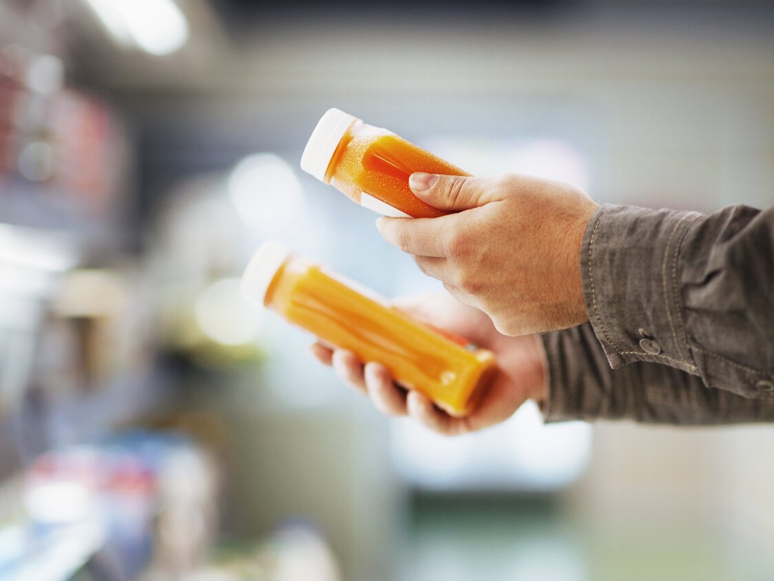  Zwei Hände, welche Smoothies halten, vor einem Kühlregal im Supermarkt.