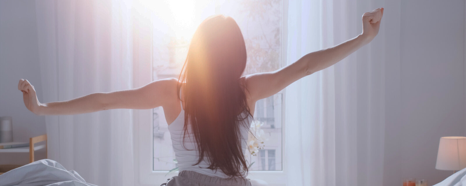 Eine Frau von hinten auf einem Bett sitzend. Sie streckt sich. Vor ihr ein Fenster, in das Licht scheint.