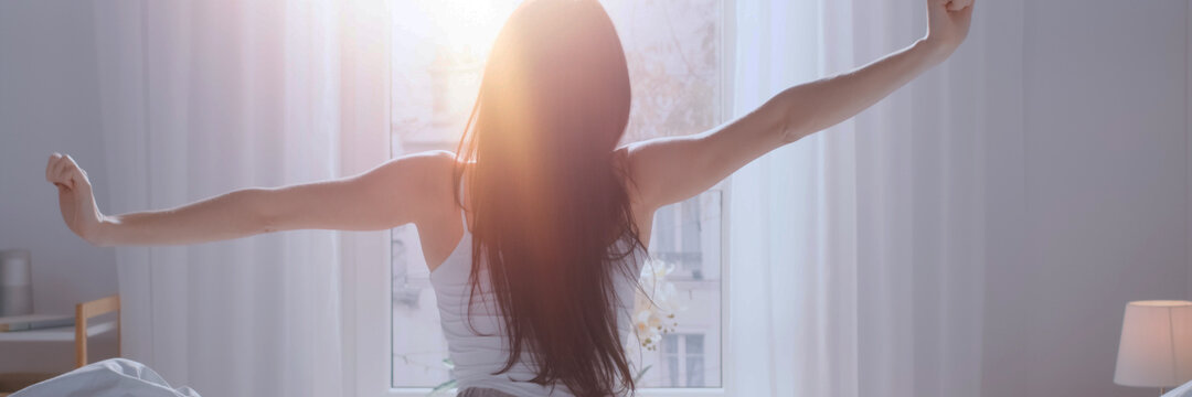 Eine Frau von hinten auf einem Bett sitzend. Sie streckt sich. Vor ihr ein Fenster, in das Licht scheint.