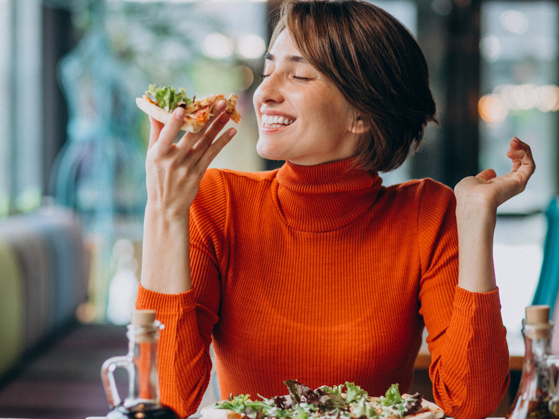 Eine Frau sitzt an einem Esstisch und isst glücklich eine Pizza.