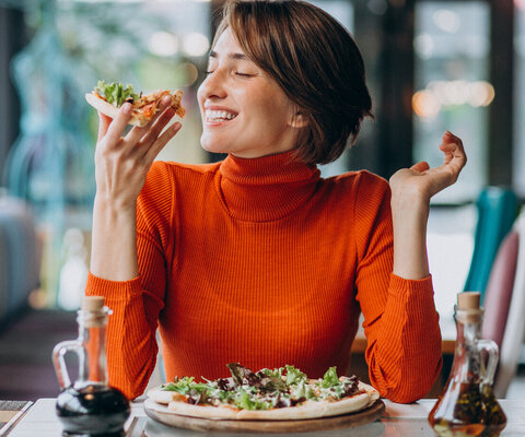 Eine Frau sitzt an einem Esstisch und isst glücklich eine Pizza.