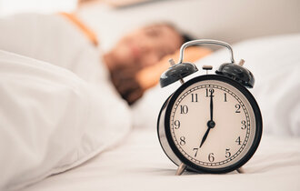 Ein großer Wecker, der 7 Uhr zeigt. Im Hintergrund unscharf eine schlafende Person.