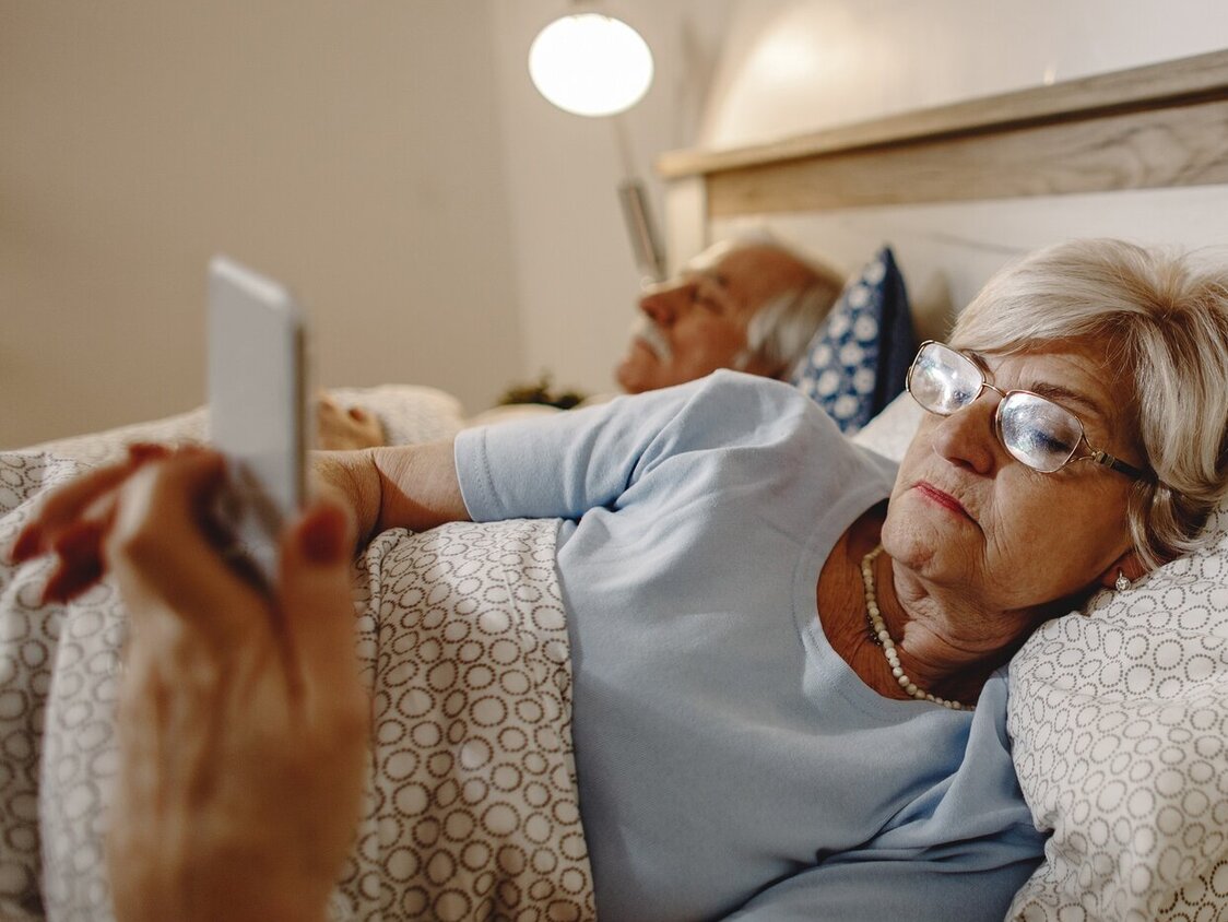 Eine ältere Frau liegt im Bett und guckt auf ihr Handy.