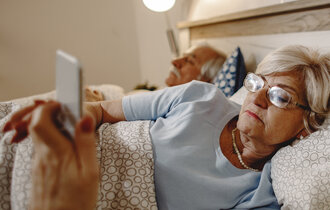 Eine ältere Frau liegt im Bett und guckt auf ihr Handy.