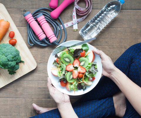 Sicht auf auf einen Teller voll gemischten Salat. Im Hintergrund eine Flasche Wasser, ein Springseil und ein Holzbrett mit einer Karotte, einem Brokkoli und einigen Cherry-Tomaten darauf.