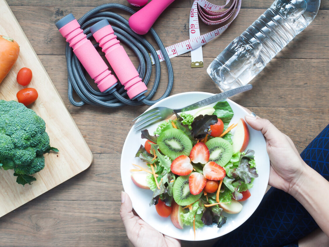 Sicht auf auf einen Teller voll gemischten Salat. Im Hintergrund eine Flasche Wasser, ein Springseil und ein Holzbrett mit einer Karotte, einem Brokkoli und einigen Cherry-Tomaten darauf.