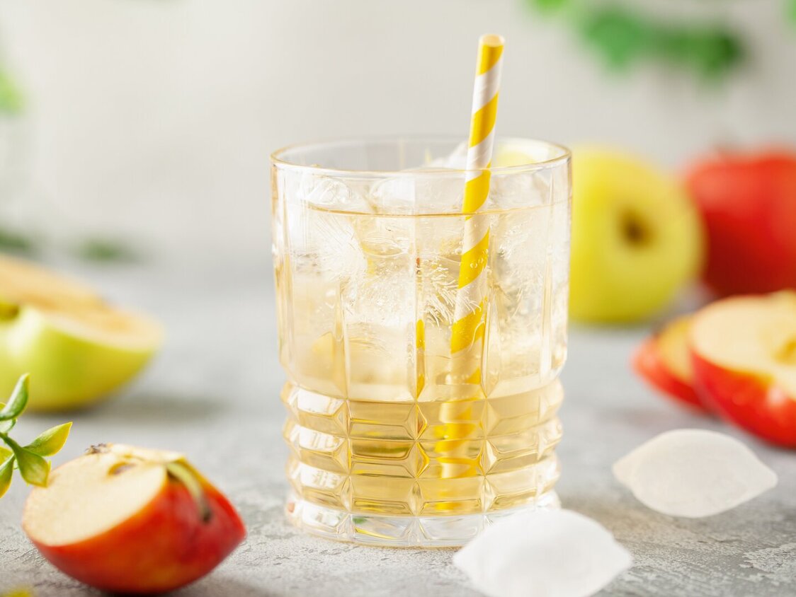 Ein Glas mit Apfelsaftschorle, Eiswürfel und ein Strohhalm und drum herum liegen Äpfel.